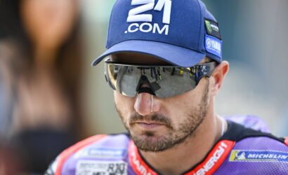 Male motorcycle racer wearing a blue cap with 21.COM and reflective sunglasses, focused on the race ahead.