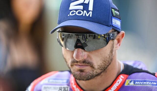 Male motorcycle racer wearing a blue cap with 21.COM and reflective sunglasses, focused on the race ahead.