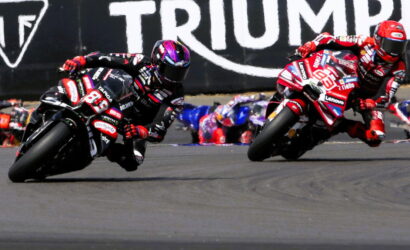 Two motorcycle racers in red and black gear lean sharply into a left-hand turn on a race track, with a Triumph banner in the background.