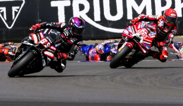 Two motorcycle racers in red and black gear lean sharply into a left-hand turn on a race track, with a Triumph banner in the background.