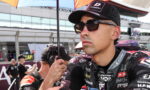 Close-up of a male motorcycle racer wearing sunglasses and a cap, standing under a pit-area umbrella.