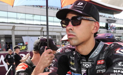 Close-up of a male motorcycle racer wearing sunglasses and a cap, standing under a pit-area umbrella.