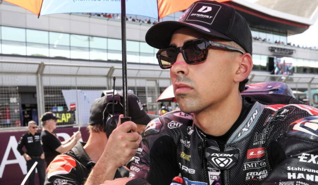 Close-up of a male motorcycle racer wearing sunglasses and a cap, standing under a pit-area umbrella.