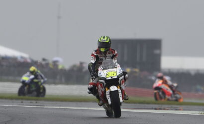 Racer in red-white suit and neon helmet on a Honda race bike #35 powers toward the camera on a wet track, with blurred riders in the background.