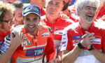MotoGP team members in red Ducati uniforms pose and smile after a race, with one rider wearing a Michelin cap in front.