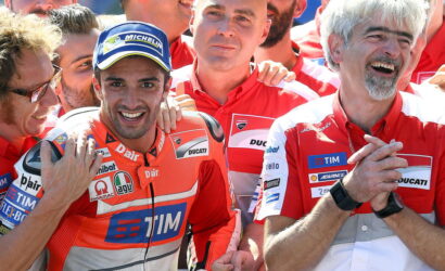 MotoGP team members in red Ducati uniforms pose and smile after a race, with one rider wearing a Michelin cap in front.