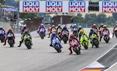 Motorcycle racers lean into a wide track start, dozens of colorful bikes racing under LIQUI MOLY banners at a circuit start line.