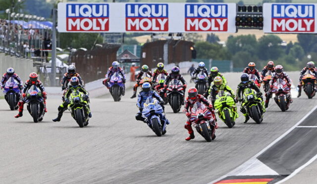 Motorcycle racers lean into a wide track start, dozens of colorful bikes racing under LIQUI MOLY banners at a circuit start line.