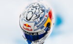 Close-up of a race helmet covered in sponsor logos (Red Bull, Heineken, Oracle) on a white shell with blue and yellow accents, blurred pale background.