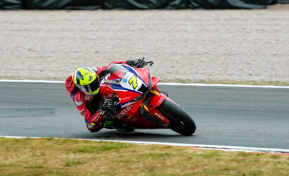 Motorcycle racer in a red Honda suit leaning into a sharp turn on a racetrack, with a bright yellow helmet and blue-white-red bike livery behind.