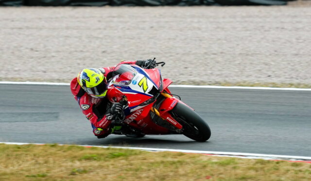 Motorcycle racer in a red Honda suit leaning into a sharp turn on a racetrack, with a bright yellow helmet and blue-white-red bike livery behind.