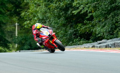 Racer in red gear leaning into a curve on a race track, yellow helmet, red motorcycle with number 7, green trees in the background.