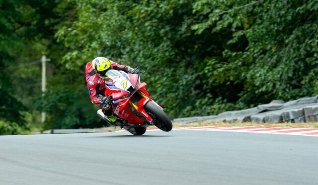 Racer in red gear leaning into a curve on a race track, yellow helmet, red motorcycle with number 7, green trees in the background.