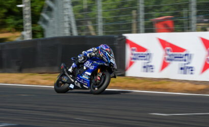 Motorcycle racer in blue riding suit leans into a turn on a blue Yamaha race bike on a racetrack; sponsor banners visible in background.