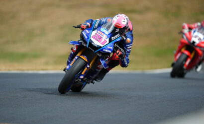 Racer in blue Yamaha suit on a blue race bike leans into a turn on a track, pink helmet visible, red bike behind them.