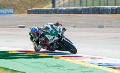 Motorcyclist in a racing suit leans into a turn on a track, colorful sponsor livery visible on the bike and suit.