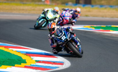 Motorcycle race: rider in blue and white leans into a sharp turn, others follow on a track with colorful curbs.