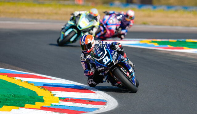 Motorcycle race: rider in blue and white leans into a sharp turn, others follow on a track with colorful curbs.