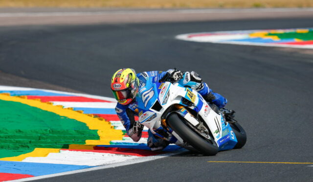Motorcycle racer in blue and white suit leaning into a sharp turn on a race track, close to colorful curb markings.