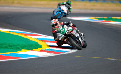 Motorcycle racer in a green-and-white suit leans into a sharp corner on a racetrack, another rider trailing behind in the background.