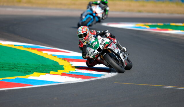 Motorcycle racer in a green-and-white suit leans into a sharp corner on a racetrack, another rider trailing behind in the background.