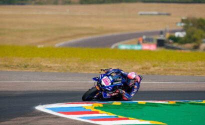 Motorcycle racer in blue gear leaning into a sharp turn on a race track, bike numbered 26 with pink helmet accents.
