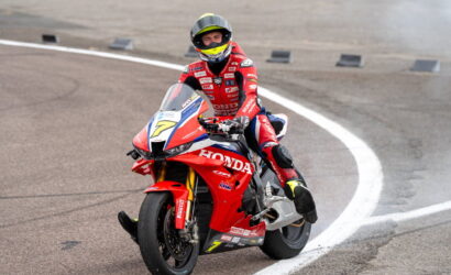 Motorcycle racer in a red Honda racing suit riding a red Honda sportbike on a track, leaning through a curve into the turn.