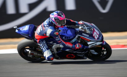 MotoGP rider in blue-red gear leaning into a turn on a blue Yamaha race bike, number 65, on a racetrack surface.