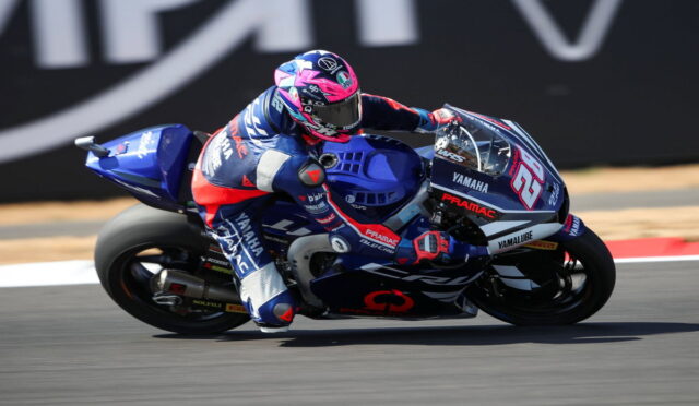 MotoGP rider in blue-red gear leaning into a turn on a blue Yamaha race bike, number 65, on a racetrack surface.