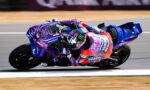 Motorcycle racer in purple Yamaha PRAMAC livery leaning into a high-speed turn on a race track, helmet with flame design.