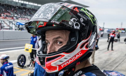 Close-up of a motorcycle racer wearing a red, white, and black helmet with sponsor logos in the pit lane behind him.