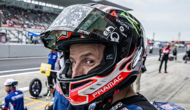 Close-up of a motorcycle racer in a red, white, and black helmet in the pit lane, looking toward the camera.
