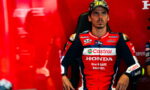 MotoGP rider in a red Honda Castrol racing suit seated in a chair against a red background in the paddock/garage area.