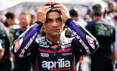 Moto rider in an Aprilia Racing suit stands in a crowded paddock, hands on his head adjusting his helmet bandage on the nose.