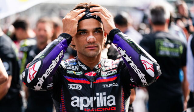 Moto rider in an Aprilia Racing suit stands in a crowded paddock, hands on his head adjusting his helmet bandage on the nose.