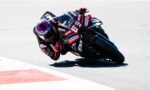 MotoGP rider in full racing gear leans into a sharp corner on a black sponsor-labeled bike, track curbing visible to the left