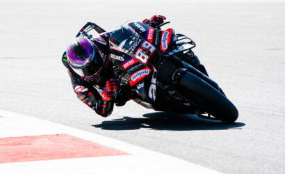 MotoGP rider in full racing gear leans into a sharp corner on a black sponsor-labeled bike, track curbing visible to the left