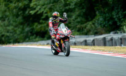 Motorcycle racer on a race track in red and black gear waves to the camera as they ride a red sportbike.