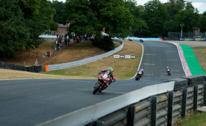 Racers lean into a sharp curve on a race track as spectators watch from a grassy hill to the left.