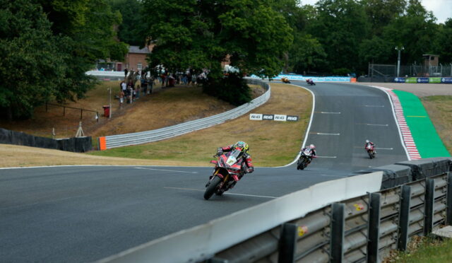 Racers lean into a sharp curve on a race track as spectators watch from a grassy hill to the left.