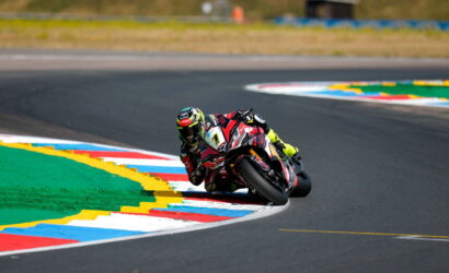Moto racer in red and black suit leans into a sharp corner on a race track with colorful curbs.