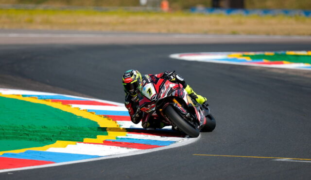 Moto racer in red and black suit leans into a sharp corner on a race track with colorful curbs.
