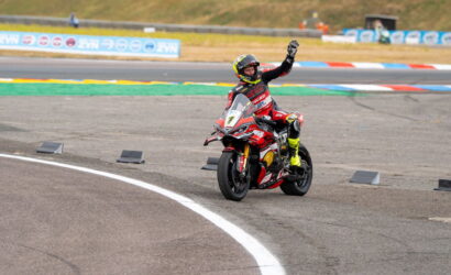 Motorcycle racer on a red Ducati sportbike on a track, helmeted rider waves as they exit a curve.