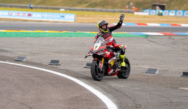 Motorcycle racer on a red Ducati sportbike on a track, helmeted rider waves as they exit a curve.