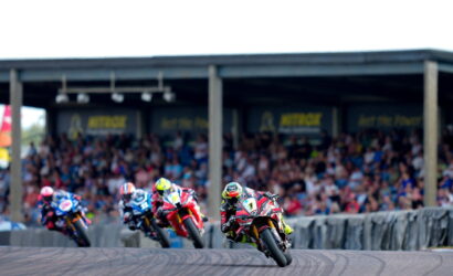 Motorcycle race on a curved track with four riders leaning into a turn as a crowded grandstand watches in the background.