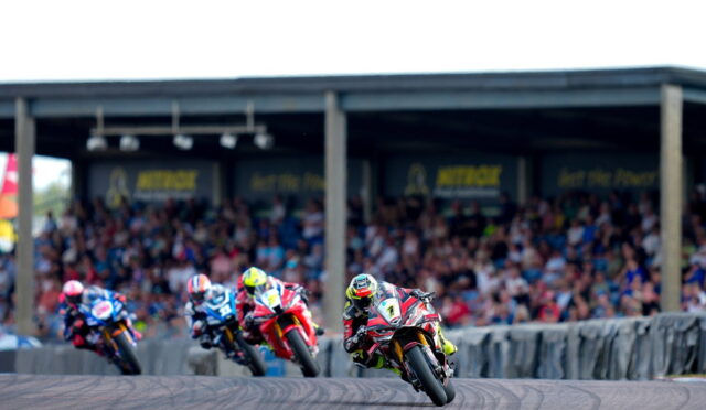 Motorcycle race on a curved track with four riders leaning into a turn as a crowded grandstand watches in the background.