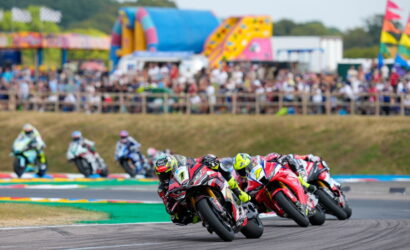 Motorcycle racers lean into a sharp curve on a track with a cheering crowd in the stands.