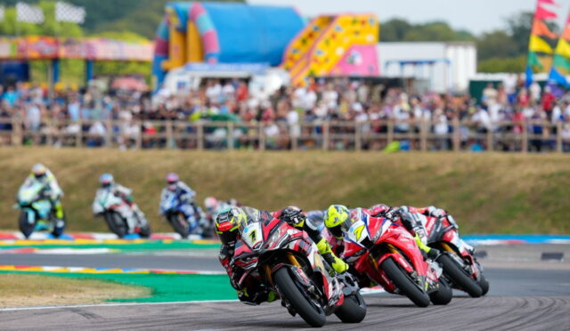 Motorcycle racers lean into a sharp curve on a track with a cheering crowd in the stands.