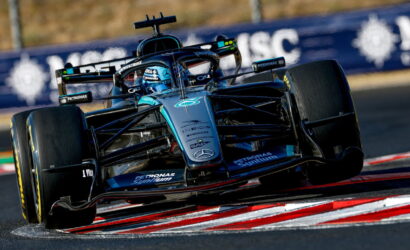 Blue Mercedes F1 race car speeding around a corner on a race track, low angle shot with red-and-white curbs visible.