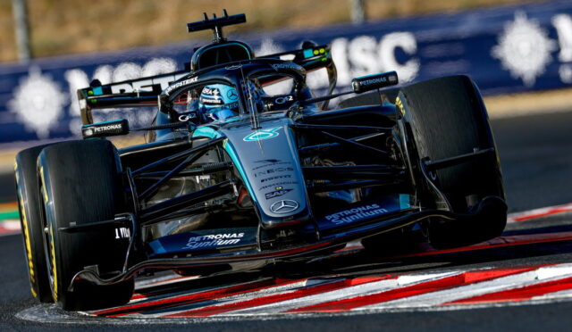 Blue Mercedes F1 race car speeding around a corner on a race track, low angle shot with red-and-white curbs visible.